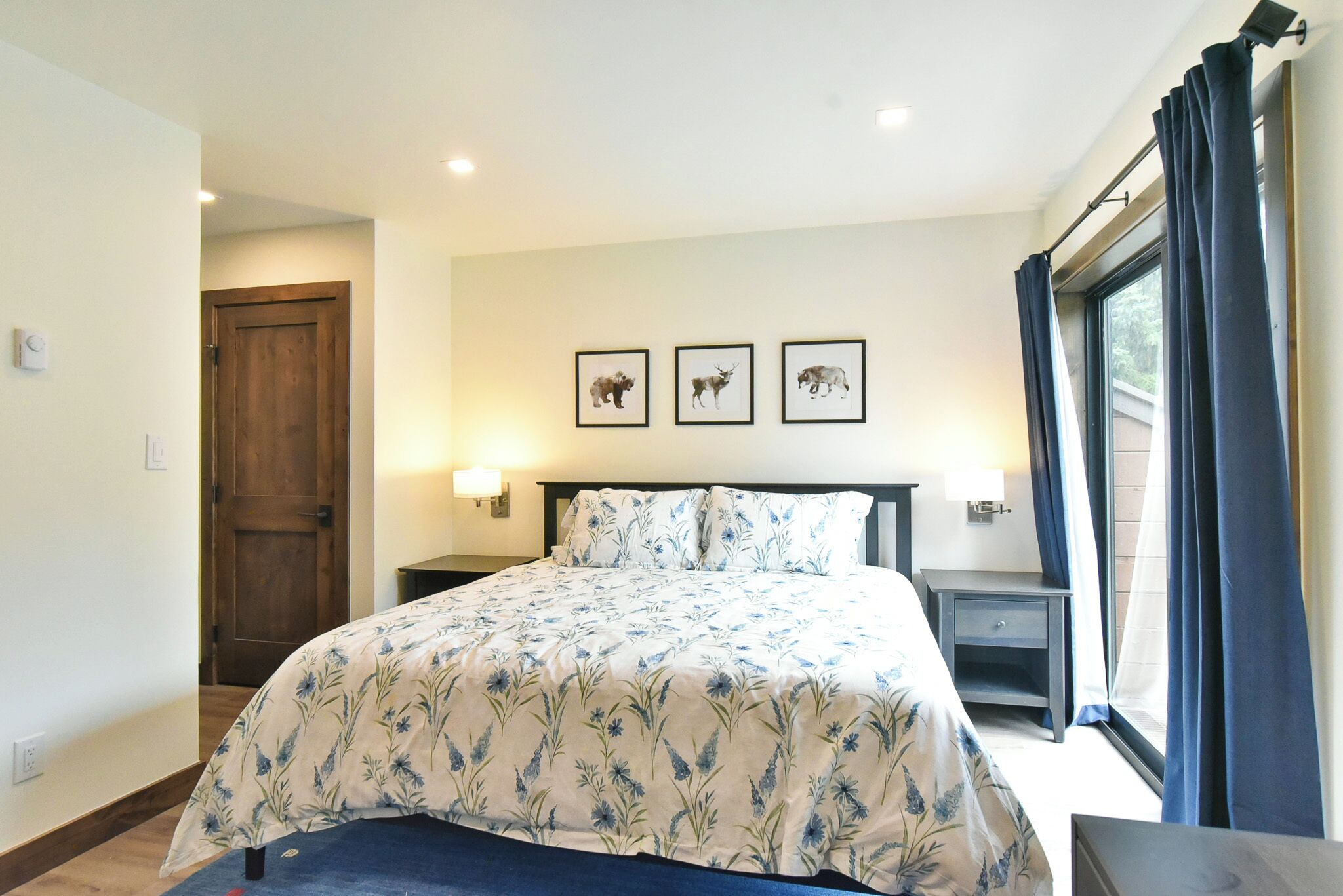 A bedroom with a bed, a nightstand, a lamp, and a window with curtains. There are three framed pictures on the wall.