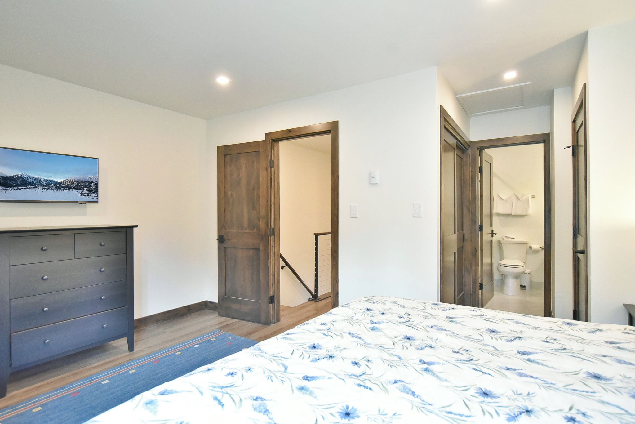 A bedroom with a bed, dresser, and a painting on the wall. There are wooden doors and a staircase visible.