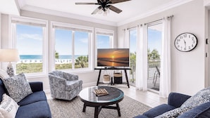 Una sala de estar con televisor, dos sofás, una mesa de centro, ventilador de techo y ventanas amplias con vista a la playa.