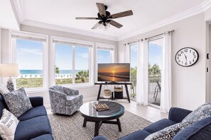 Een woonkamer met een televisie, twee banken, een salontafel, een plafondventilator en grote ramen met uitzicht op het strand.