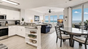 Una cocina moderna con armarios blancos, un comedor con mesa de vidrio y una sala de estar con vista al océano.