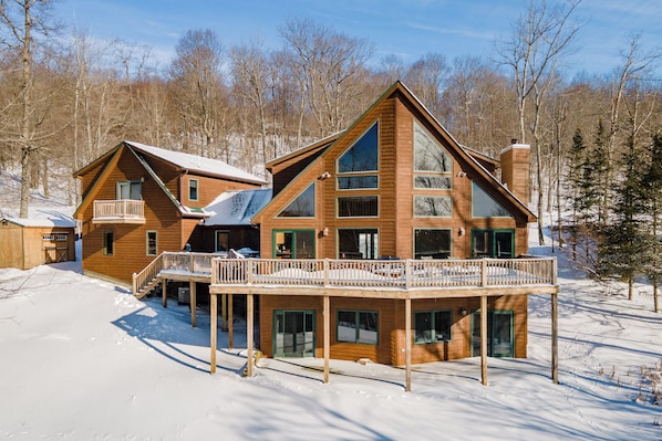 A large wooden cabin with a balcony and multiple windows, surrounded by a snowy landscape and trees.