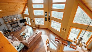 A spacious living room with a stone fireplace, large windows, and a high ceiling with wooden beams. - Mountain Chalet w/ Hot Tub, Views & Game Room (McHenry)