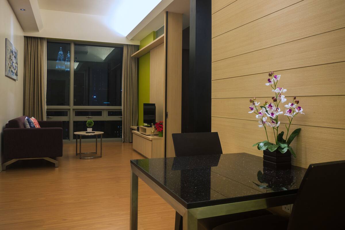 A modern living room with a glass dining table, a sofa, and a large window with a view of a cityscape at night.