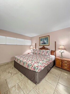 A bedroom with a bed, two bedside tables, a nightstand, a wall-mounted shelf, and a window with blinds.