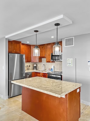 Una cocina moderna con refrigerador de acero inoxidable, encimera de granito, armarios de madera e iluminación colgante.