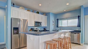 A kitchen with white cabinets, a stainless steel refrigerator, a microwave, a sink, and wooden bar stools.