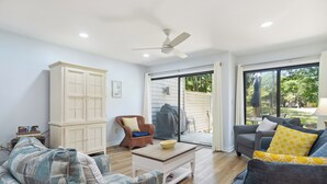 Un salon spacieux doté d’une grande fenêtre, d’un ventilateur de plafond et d’une porte-fenêtre coulissante donnant sur un espace extérieur.