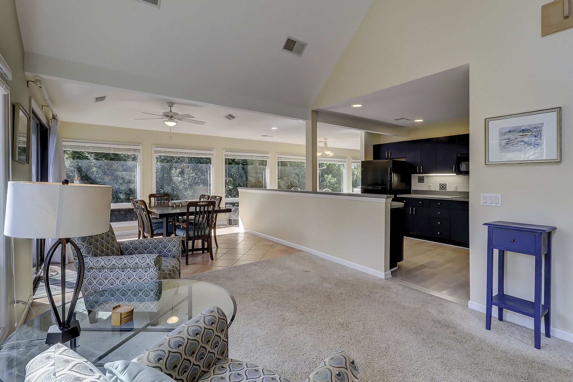A spacious living area with a dining table, comfortable seating, a kitchen with dark cabinets, and large windows offering a view of greenery.