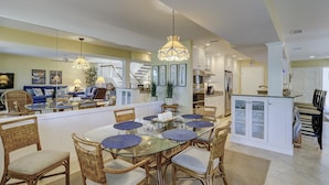 Une cuisine spacieuse dotée d’un îlot central, un coin repas avec des chaises en osier et une table en verre, et un salon avec un canapé bleu.