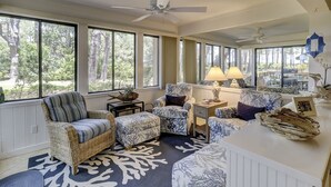 Une pièce lumineuse, baignée de soleil, avec de grandes fenêtres, des meubles en osier, un tapis à motifs et un ventilateur de plafond.