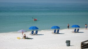 A beach with white sand, clear blue water, and several blue umbrellas providing shade.