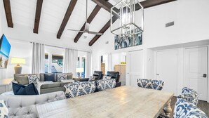 A spacious living room with a large wooden dining table, comfortable seating, and a high ceiling with exposed beams.