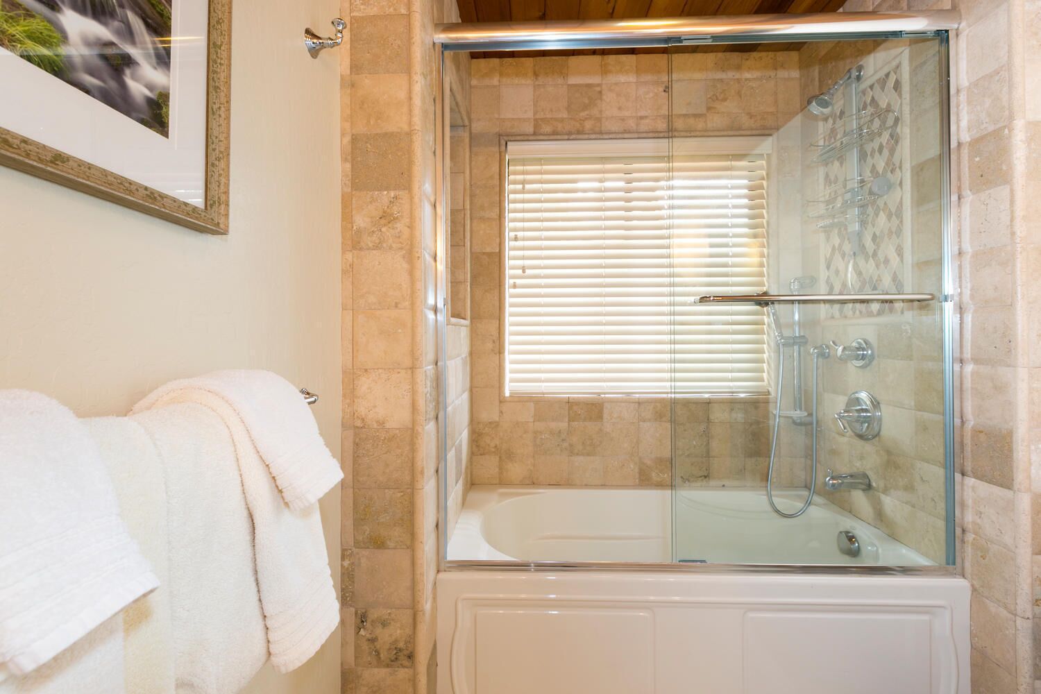 Eine gläserne Dusche mit Badewanne, beigefarbene Fliesen und einem gerahmten Bild an der Wand.