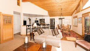 Amplia sala de estar con techo alto y vigas de madera, un sofá cómodo, un comedor con sillas de madera y una cocina al fondo.