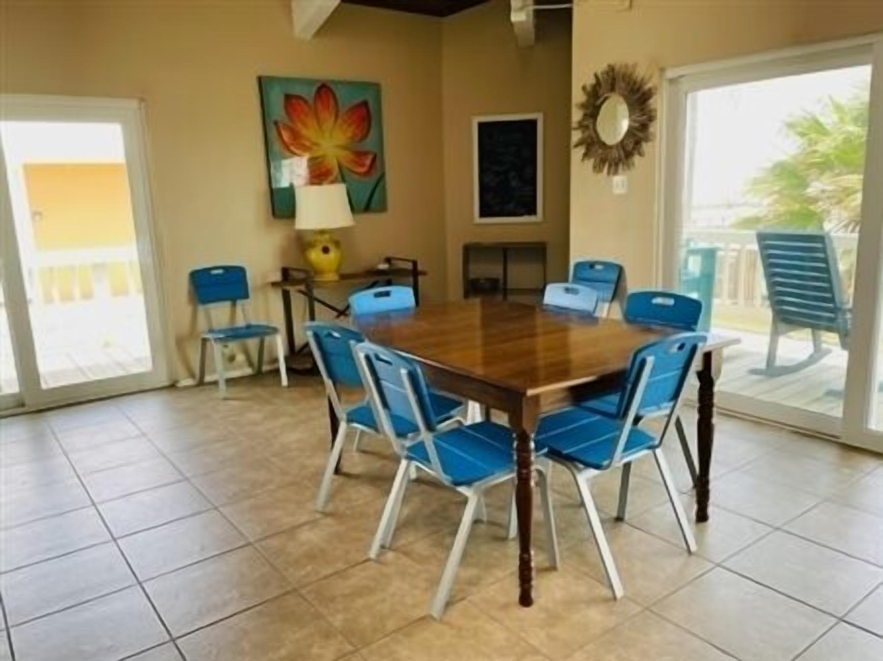 A dining area with a wooden table and blue chairs, a painting on the wall, and a view through large glass doors.
