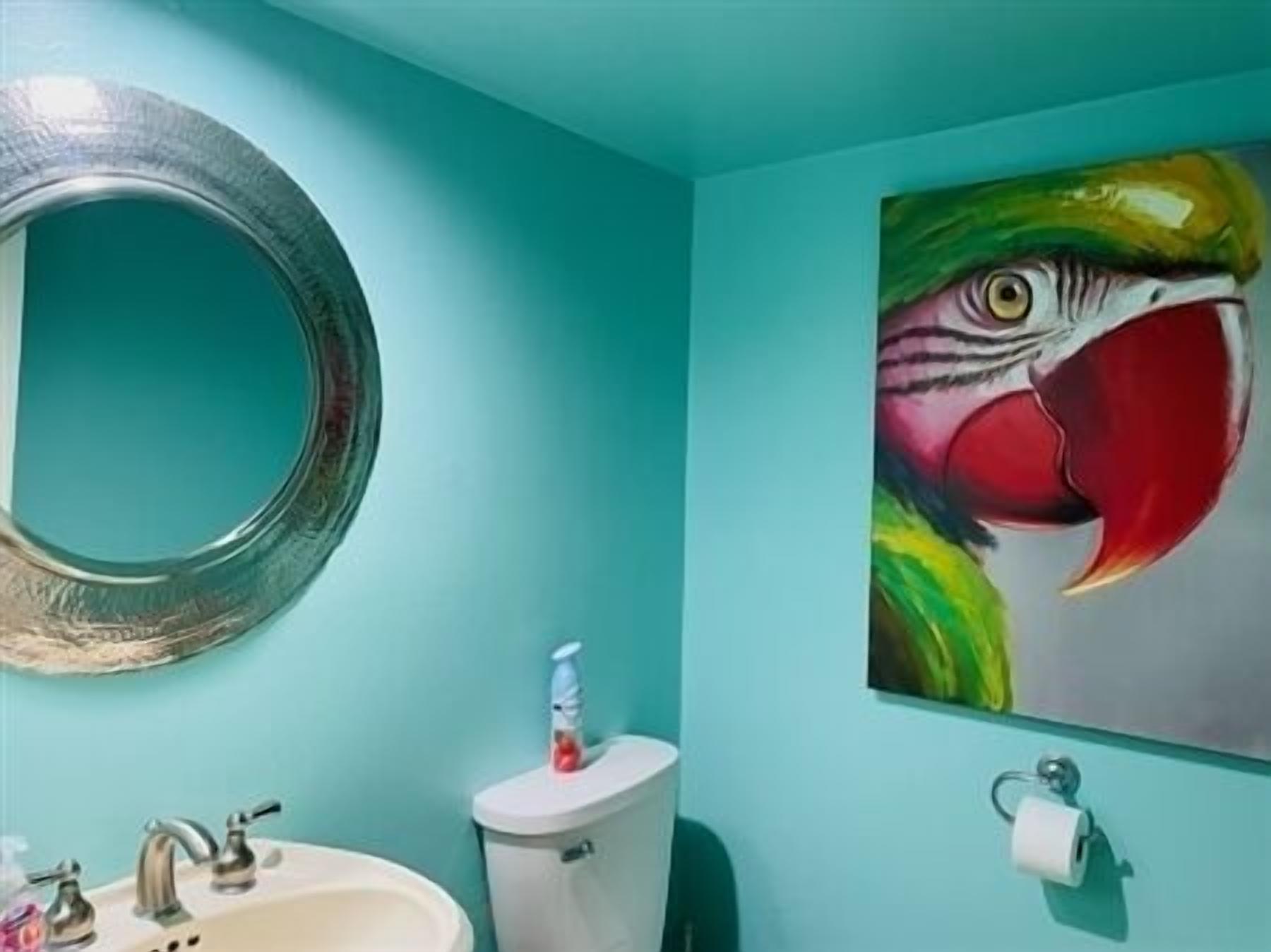 A bathroom with a teal wall, a round mirror, a toilet, a sink, and a colorful parrot painting.