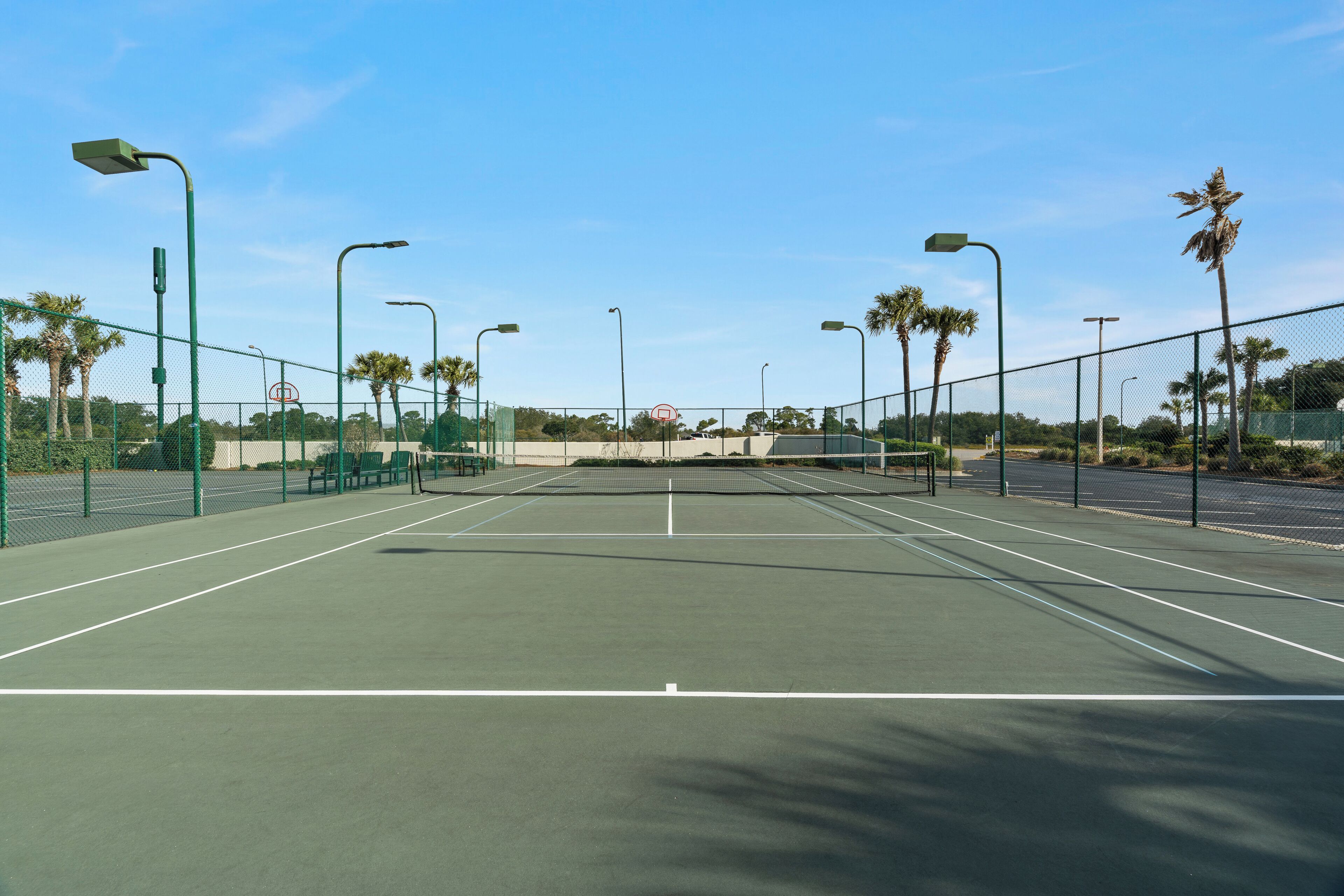 Een tennisbaan met groene hekken, palmbomen en lantaarns.