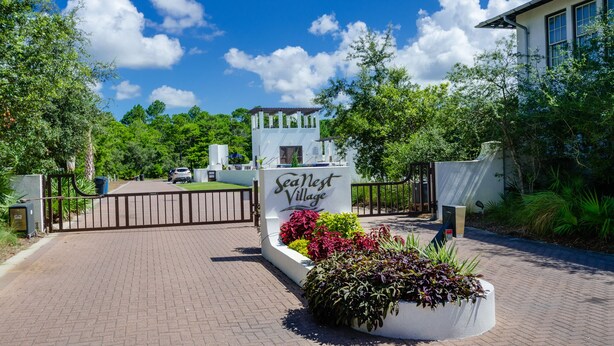 A gated entrance with a sign reading "SeaNest Village" surrounded by lush greenery and a clear blue sky.