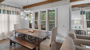 Un coin repas avec une table en bois, des chaises et un banc. De grandes fenêtres sont agrémentées de rideaux, un luminaire suspendu éclaire l’espace, et une petite table avec une lampe est également présente.