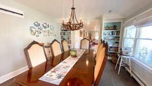 A dining room with a long wooden table, six chairs, a chandelier, and a view of a library with bookshelves.