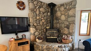 A cozy living room with a stone fireplace, a flat-screen TV, wooden furniture, and a large window.