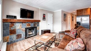 Una sala de estar con chimenea, televisor y una zona de cocina con refrigerador.