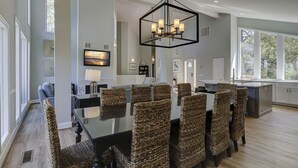 Uma sala de jantar espaçosa com uma mesa de vidro rodeada por cadeiras de fibra natural, um lustre moderno e vista para a cozinha e sala de jantar.
