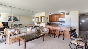 A living room with a sofa, coffee table, and chairs, a kitchen with wooden cabinets and appliances, and a glass partition. - Oceanfront Oasis: Ground Floor Retreat - Sugar Beach #130 (Kihei, Maui)