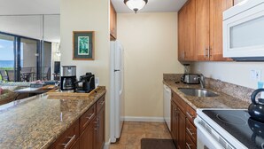 A kitchen with granite countertops, wooden cabinets, a refrigerator, a microwave, a stove, and a coffee maker. - Oceanfront Oasis: Ground Floor Retreat - Sugar Beach #130 (Kihei, Maui)