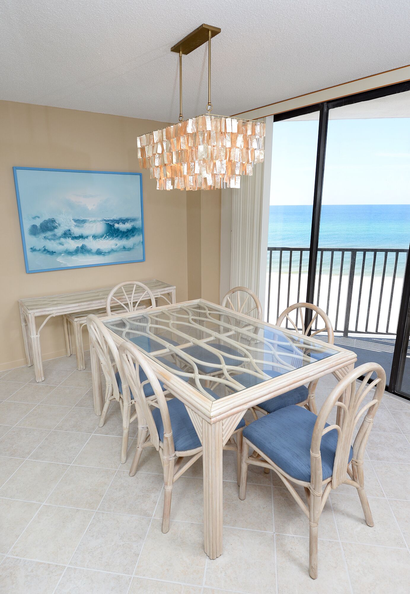 A dining area with a glass-top table and wicker chairs, a large painting of a seascape, and a view of the ocean through a balcony.