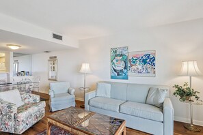 Una sala de estar con un sofá azul claro, un sillón estampado, una mesa de centro de vidrio y dos lámparas de pie. Hay dos cuadros enmarcados en la pared.