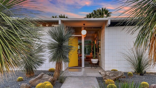 Une maison moderne avec une porte jaune, un chemin de pierres et divers cactus et plantes succulentes dans le jardin.