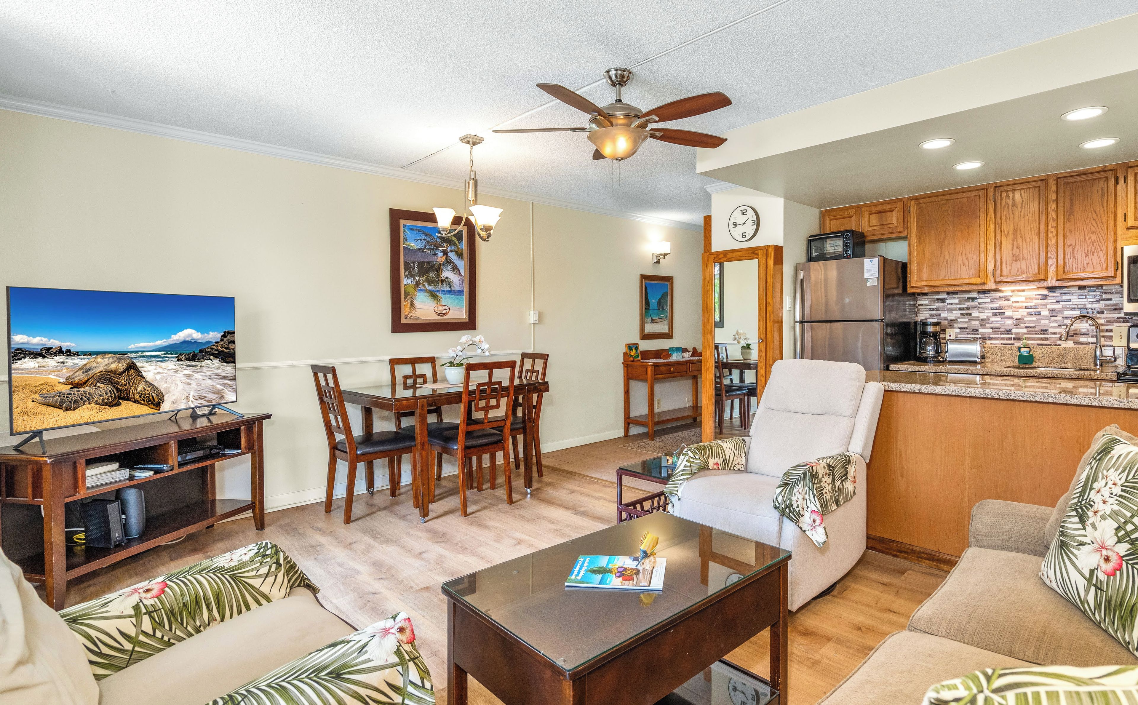 A living room with a TV, wooden furniture, a ceiling fan, and a kitchen with wooden cabinets and a refrigerator.