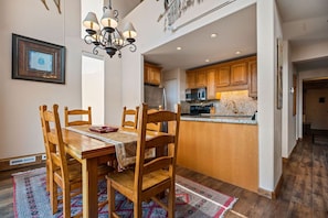 A dining area with a wooden table and chairs, a chandelier, and a kitchen in the background.