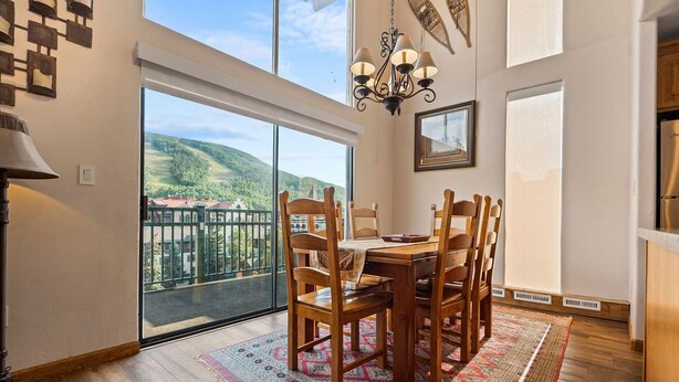 A dining area with a large window, wooden chairs, a chandelier, and a patterned rug. - Lionshead Village Condo With Mountain Views, Hot Tubs, Short Walk to Gondola (Vail)