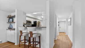 Una cocina moderna con armarios blancos, un microondas y un refrigerador. Hay dos taburetes de bar con patas de madera y asientos acolchados. El piso es de madera y las paredes son blancas.