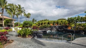 A resort pool area with a clear view of the pool, surrounding tropical vegetation, and a building with a balcony. - Summer Special! Discover luxury at Leilani Kai, a 3BR condo in beautiful Waikoloa, HI. (Waikoloa Village)