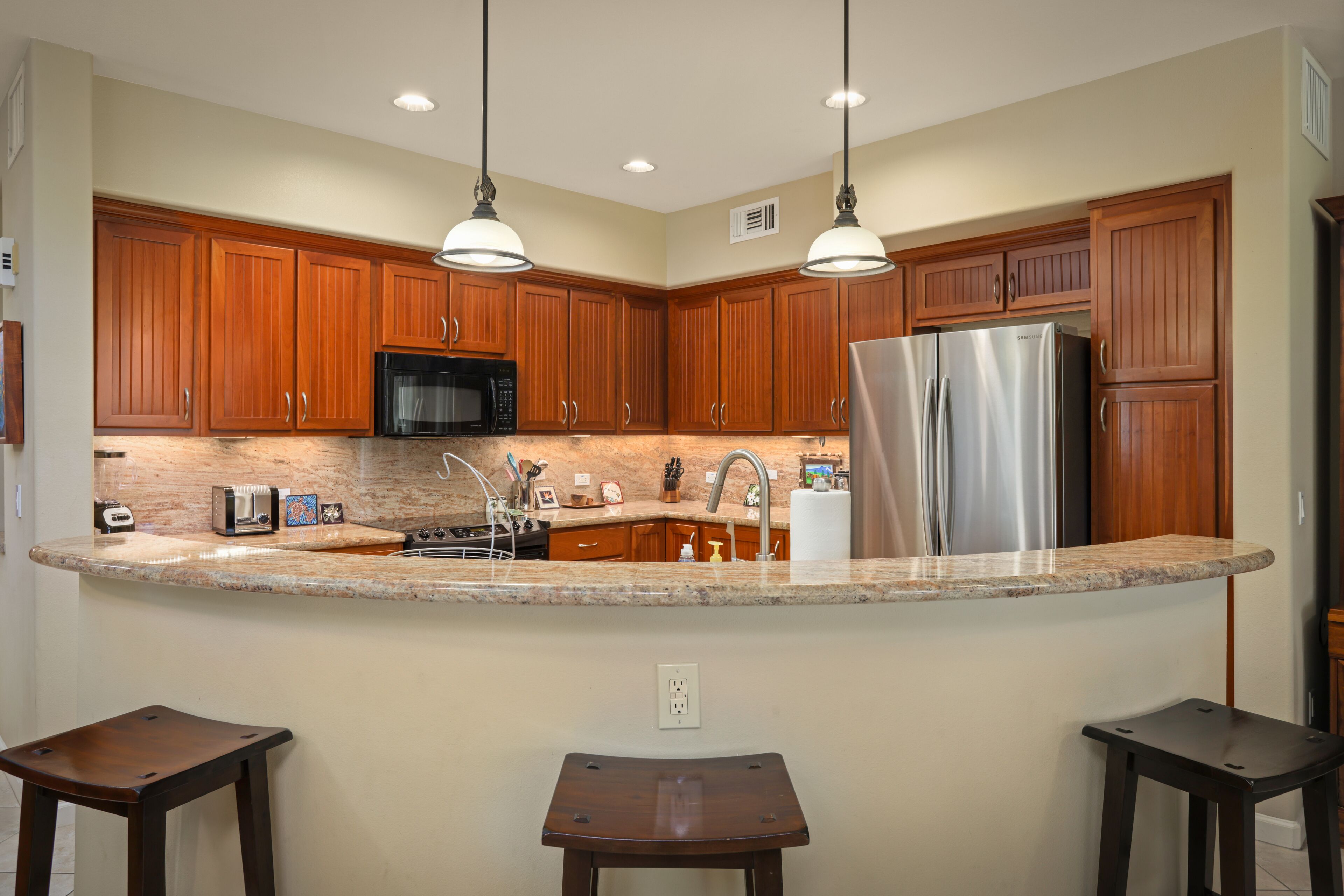 A kitchen with wooden cabinets, a stainless steel refrigerator, a microwave, and a granite countertop. There are two bar stools and pendant lights above the counter.
