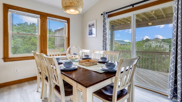 Uma sala de jantar com uma mesa de madeira posta para quatro pessoas, cercada por cadeiras brancas, janelas amplas e vista para áreas verdes e casas.