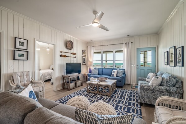 A spacious living room with a ceiling fan, a blue sofa, a patterned rug, and a view of the ocean through large windows.