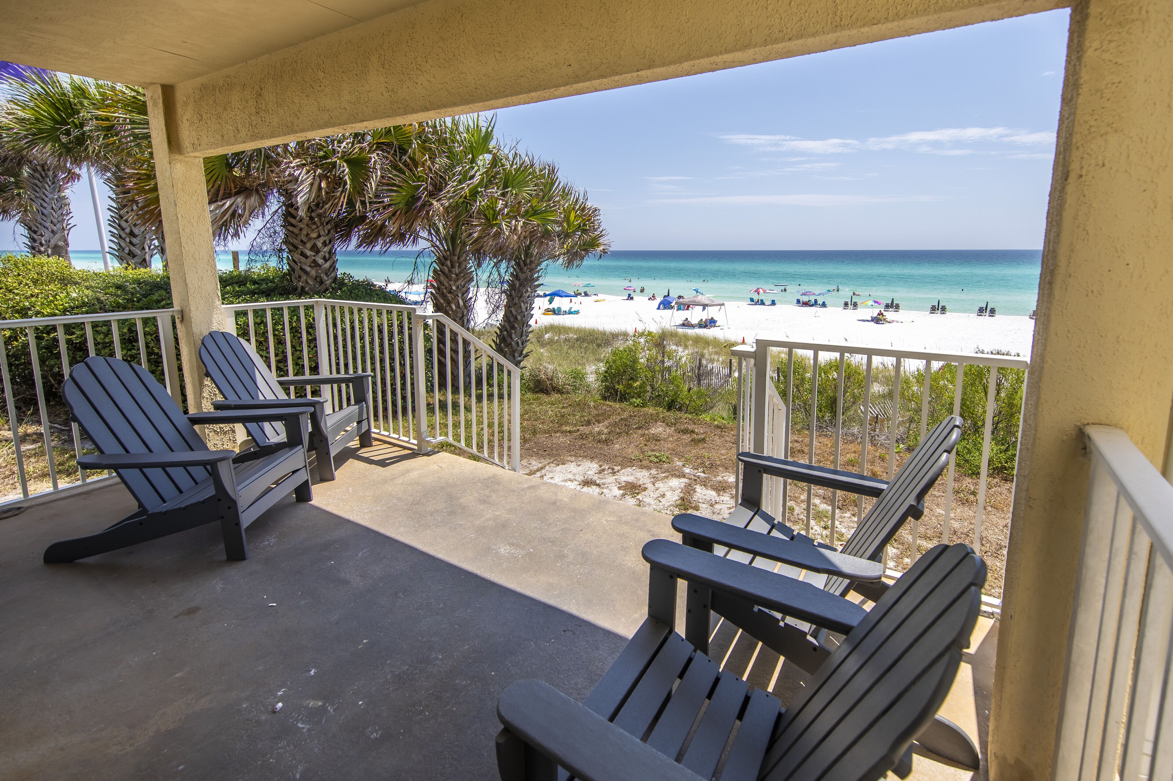 Un balcon avec des fauteuils Adirondack donnant sur une plage parsemée de parasols et où se trouvent des gens.