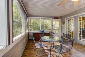 Relax in this serene sunroom with a glass table and metal chairs, surrounded by large windows offering a view of the lush trees.