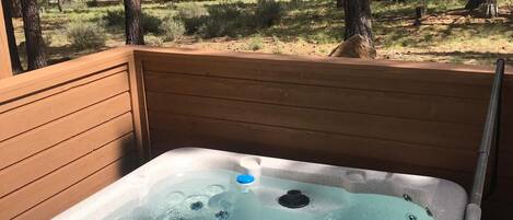 A hot tub with a control panel, surrounded by a wooden deck and a forest view.