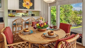 A dining area with a round wooden table, wicker chairs, and a large window offering a view of greenery. - Manualoha 1105 Ocean Mountain views By Suite Paradise (Koloa)