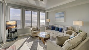 Een ruime woonkamer met een groot raam met uitzicht op het strand, een comfortabele zithoek, een glazen salontafel en een flatscreen-tv.