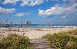 A beach with a wooden boardwalk leading to the water, clear skies, and sparse vegetation.
