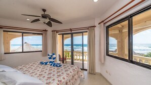 Une chambre d’hôtel avec un lit, un ventilateur de plafond, de grandes fenêtres donnant sur l’océan et un balcon avec une balustrade.
