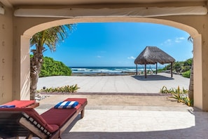 A beach view from a covered area with a lounge chair and a thatched structure.