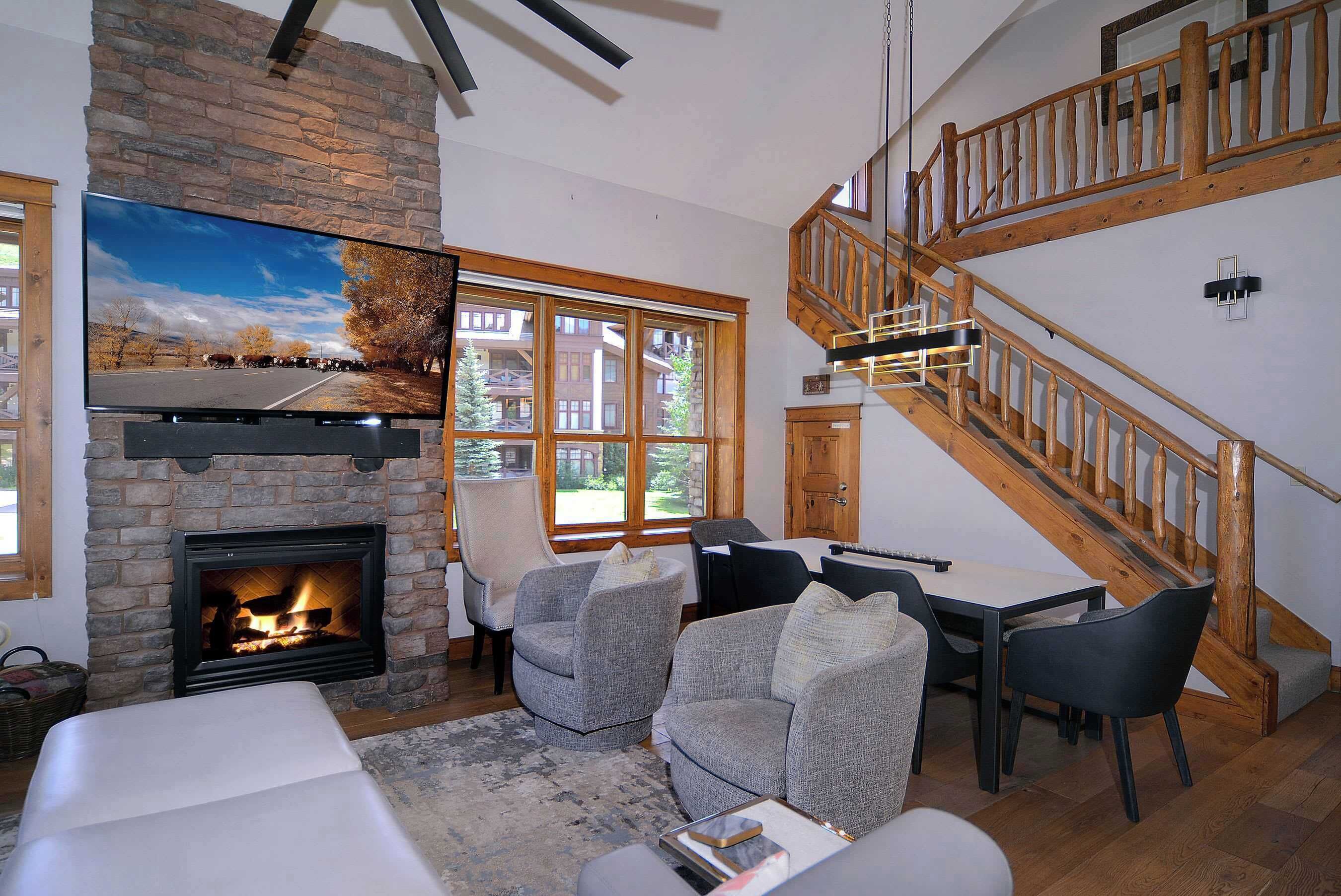 A living room with a stone fireplace, a television, a staircase, and a view of a road and trees.